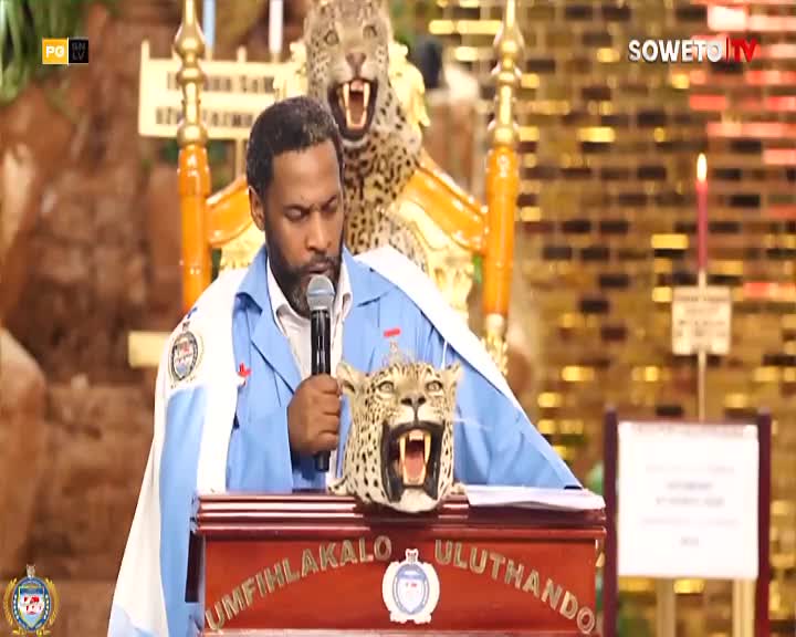 A man in a light blue shirt and a white and blue cape stands at a podium, speaking into a microphone. Behind him, a stuffed leopard head rests on the podium, and another leopard skin is draped over a golden throne. The Soweto TV logo is visible in the upper right corner.
