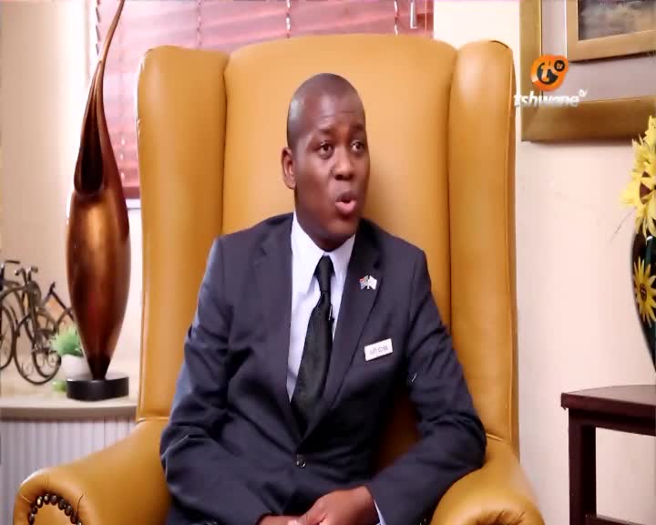 The man in the suit sits in a large yellow armchair, speaking animatedly. A Tshwane TV logo is visible in the corner, suggesting a South African interview.
