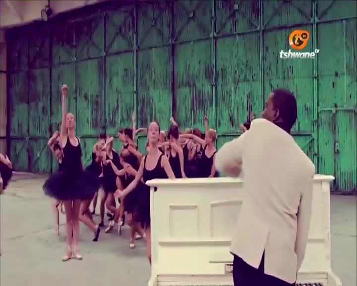 A group of dancers in black leotards and tutus are moving gracefully, arms raised, in front of a white piano. The man in the white jacket, likely the conductor, gestures with his hands as the performance unfolds, possibly for Tshwane TV in South Africa.

