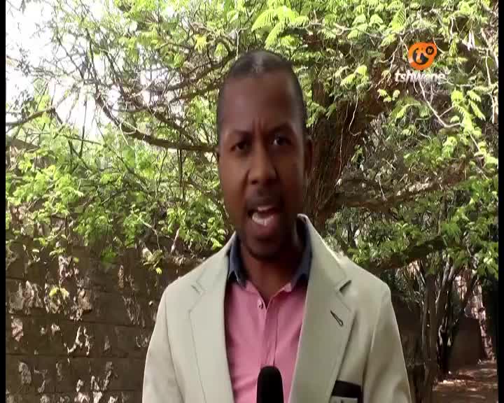 A man in a blazer speaks directly into a microphone, his mouth open as if mid-sentence. Behind him, the Tshwane TV logo is visible, with greenery and a brick wall providing a backdrop.
