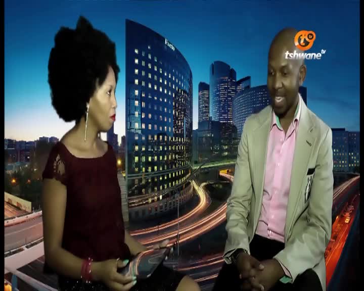 The man in the blazer is speaking, his hands clasped in front of him, while the woman in the burgundy top looks on. The Tshwane TV logo is visible, and the backdrop shows a cityscape with bright lights.
