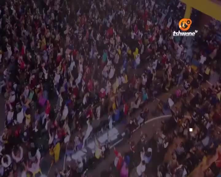 A sea of people, mostly in white and red, are packed together, their arms raised. The Tshwane TV logo is visible, broadcasting the event to South Africa.
