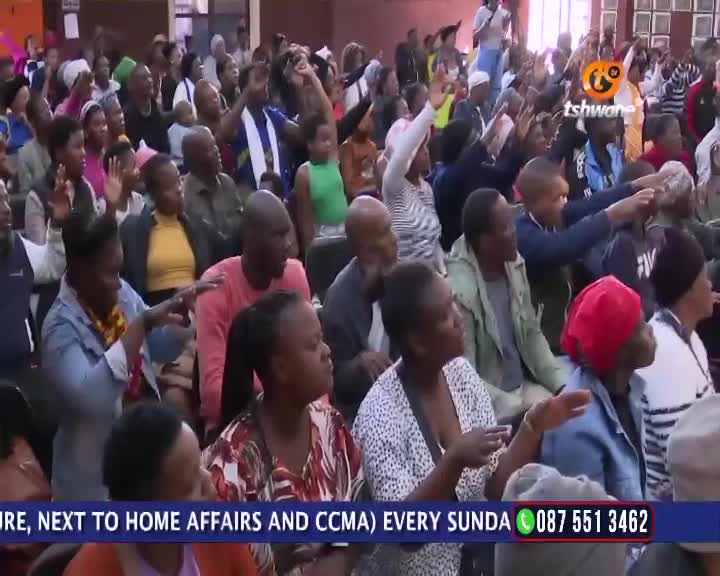 People in the crowd raise their hands, some pointing toward the front. A Tshwane TV logo is visible, and a phone number is displayed at the bottom of the screen.
