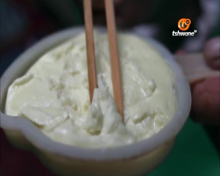 A pair of wooden chopsticks are gently stirring a creamy, pale yellow mixture in a small bowl. The Tshwane TV logo is visible in the corner, suggesting this is a cooking segment.
