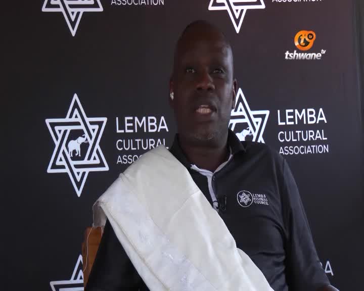 A man in a black shirt and white shawl is speaking, likely for Tshwane TV, with a Lemba Cultural Association backdrop. His expression is serious as he addresses the camera.

