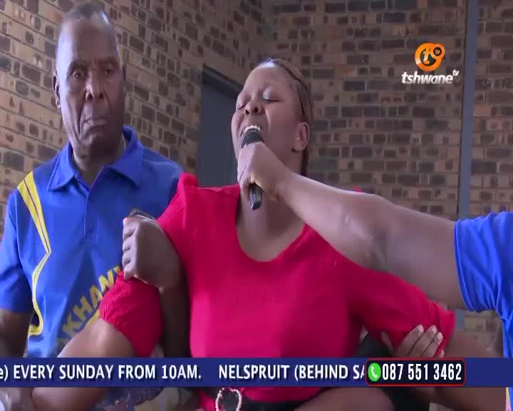 A woman in a red shirt sings into a microphone, her arms held by others on either side. Behind her, a man in a blue shirt looks on, the Tshwane TV logo visible in the corner.
