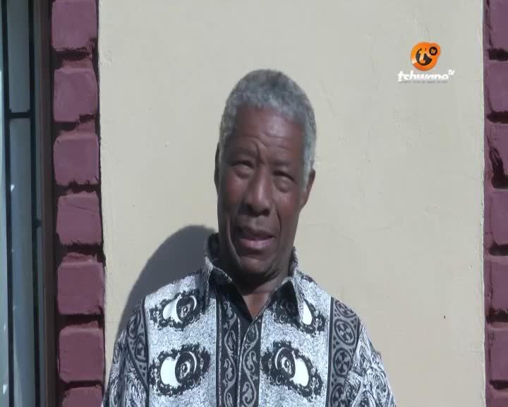 A man in a patterned shirt is speaking, his face lit by the sun. The Tshwane TV logo sits above his head, and a brick wall is visible to his right.
