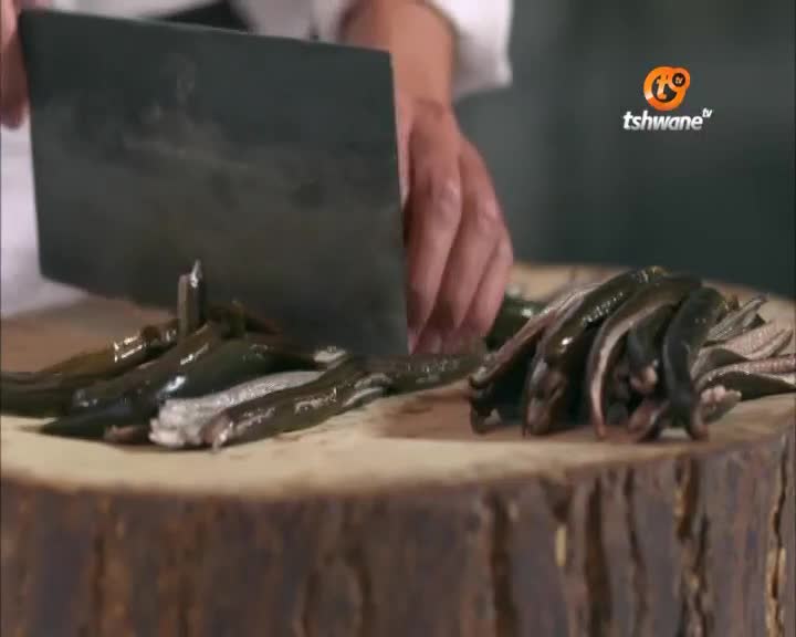 A large cleaver descends, chopping dark, slender fish on a wooden cutting board. The Tshwane TV logo sits in the corner as the chef prepares the meal.
