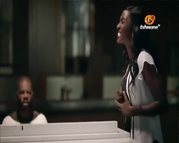 A woman in a white blouse is singing, her face contorted with emotion, as a man sits at a piano on Tshwane TV. The scene is dimly lit, and the focus is on the singer's passionate performance.
