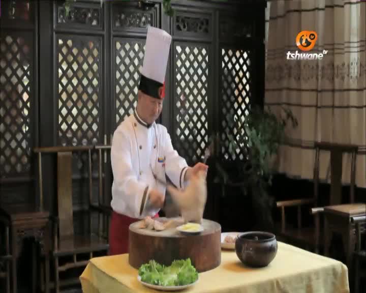 A chef in a tall white hat and jacket is working at a table, preparing food for a Tshwane TV segment. He's holding a piece of meat over a wooden cutting board, with plates of ingredients arranged nearby.
A chef in a tall white hat and jacket is working at a table, preparing food for a Tshwane TV segment. He's holding a piece of meat over a wooden cutting board, with plates of ingredients arranged nearby.