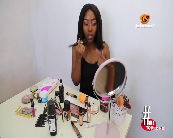 A woman, framed by a Tshwane TV logo, holds a brush to her cheek, applying makeup. Around her, a table is covered with products, a mirror reflecting her focused expression.

