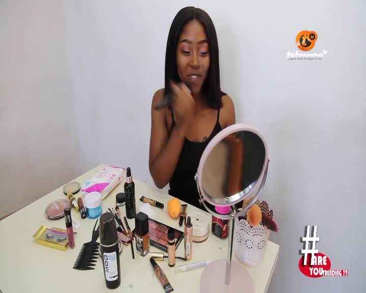 She's holding a brush, sweeping it across her face as she looks into the mirror. The Tshwane TV logo is in the background, and the table is covered in makeup.
