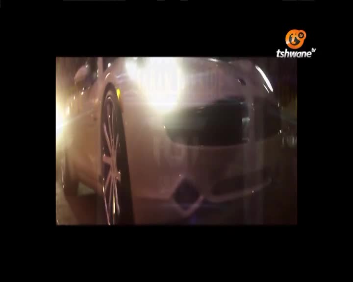 A bright car is approaching, its headlights blinding in the dark. The Tshwane TV logo is in the corner as the vehicle's chrome wheels gleam.
