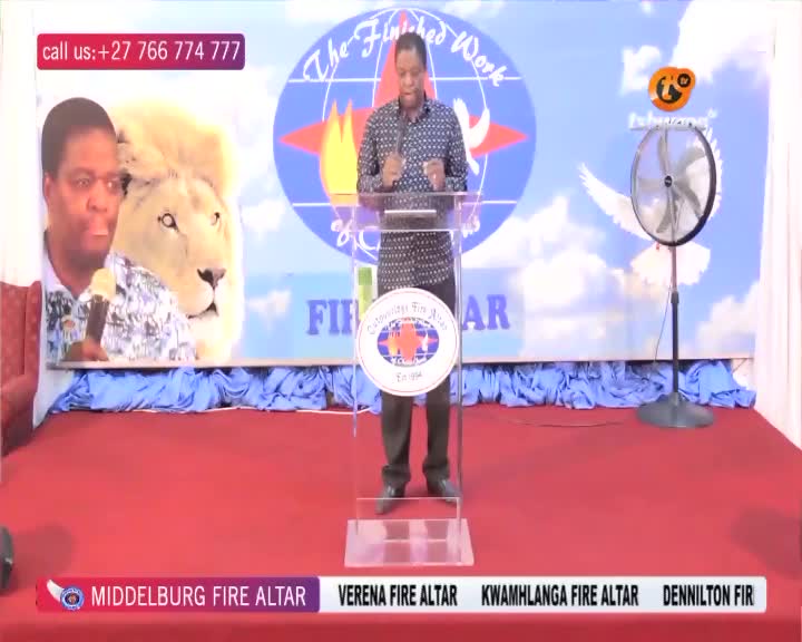 A man stands behind a clear podium, speaking into a microphone. Behind him, a large banner displays a lion's head and the words "The Finished Work." A fan spins lazily to the right.