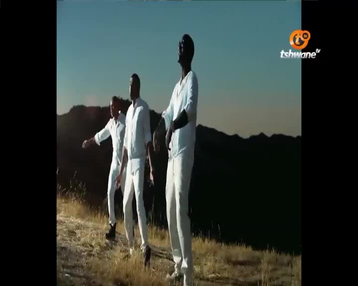 Three men in white outfits move their arms in unison against a backdrop of rolling hills under a clear sky. The Tshwane TV logo appears in the upper right corner.