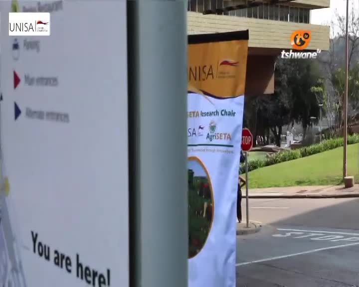 A person in a dark shirt walks past a banner advertising the UNISA Agriseta Research Chair. The Tshwane TV logo is visible on the building behind them.
