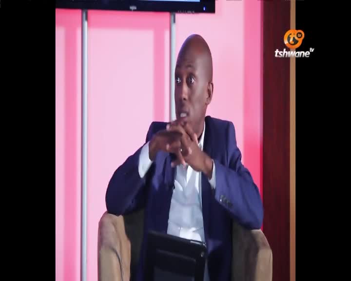 A man in a blue suit leans forward, his hands clasped in front of him. He's speaking on a program for Tshwane TV.