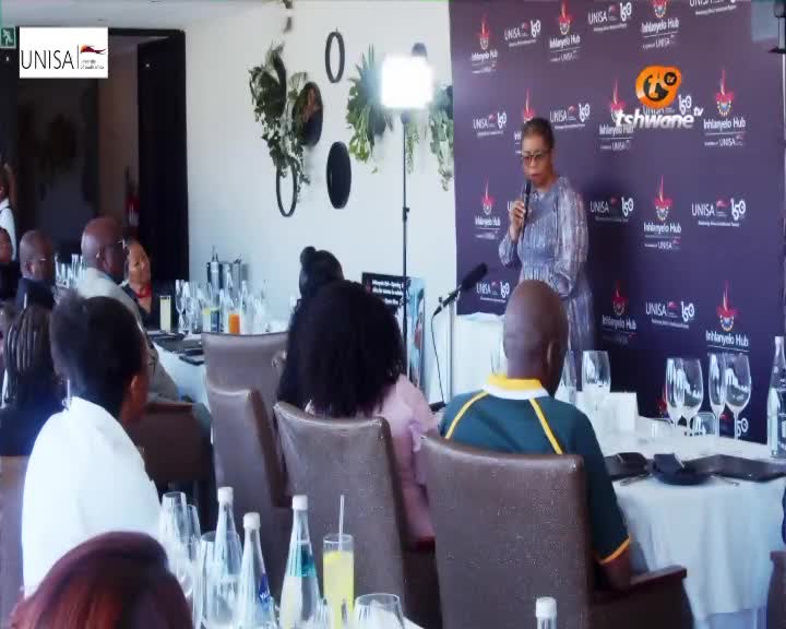 A woman stands at a podium, speaking into a microphone. The backdrop behind her features the Tshwane TV logo and UNISA branding. A woman stands at a podium, speaking into a microphone. The backdrop behind her features the Tshwane TV logo and UNISA branding.
