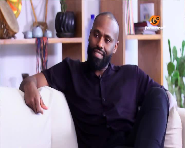 A man with a short beard and bald head, wearing a dark purple shirt, leans back on a white couch. Behind him, shelves hold decorative items and a plant, with the Tshwane TV logo visible. A man with a short beard and bald head, wearing a dark purple shirt, leans back on a white couch. Behind him, shelves hold decorative items and a plant, with the Tshwane TV logo visible.