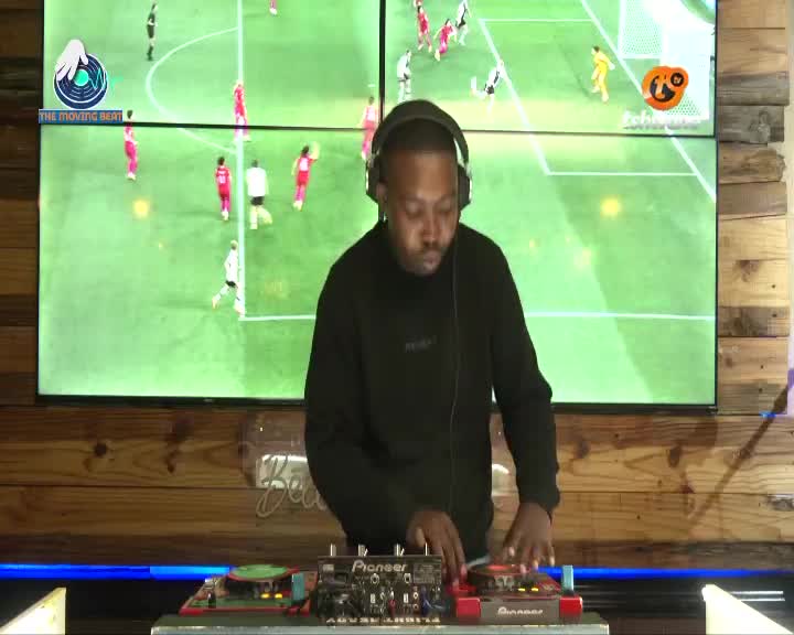 The DJ, wearing headphones, focuses on the Pioneer equipment in front of him. Behind him, two large screens display a soccer match, with players in red and white kits on a green field. The DJ, wearing headphones, focuses on the Pioneer equipment in front of him. Behind him, two large screens display a soccer match, with players in red and white kits on a green field.