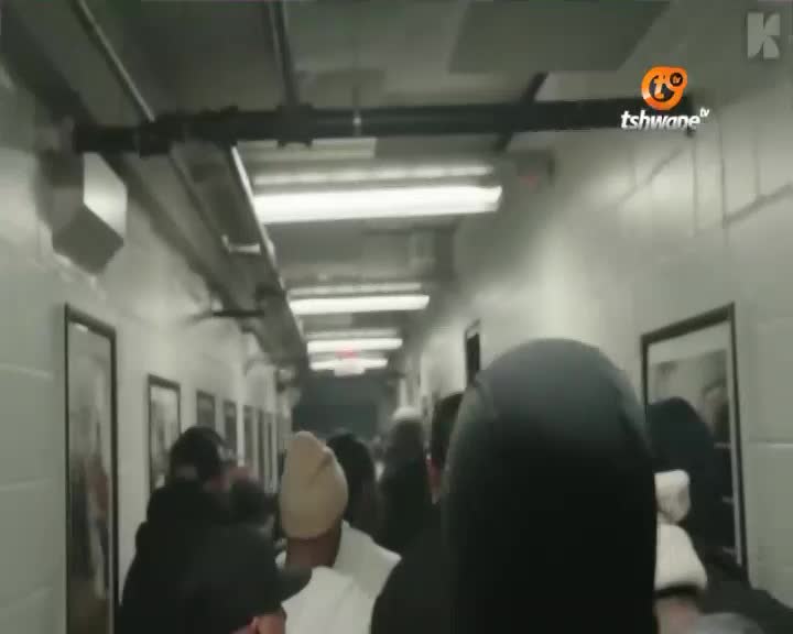 A crowd of people moves down a brightly lit corridor, the Tshwane TV logo visible on the wall. Framed pictures line the white brick walls as the group heads deeper into the building. A crowd of people moves down a brightly lit corridor, the Tshwane TV logo visible on the wall. Framed pictures line the white brick walls as the group heads deeper into the building.