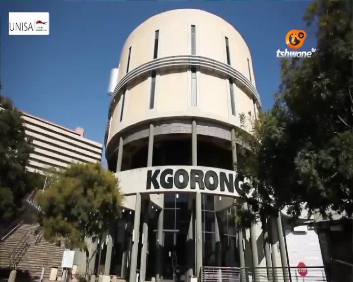 The KGORONG building, part of UNISA in Tshwane, stands tall against a clear blue sky. Lush green trees frame the entrance, their leaves rustling gently.