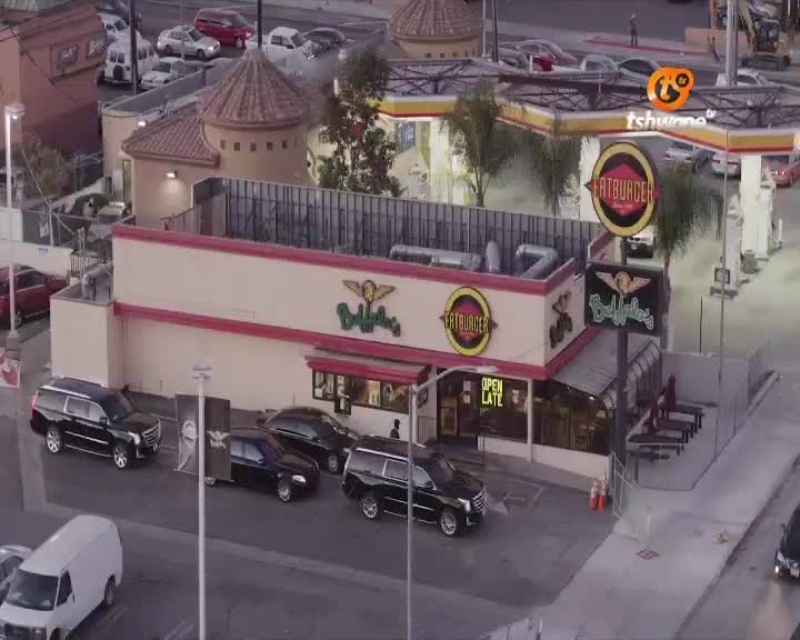 Several black SUVs and sedans are parked outside a Fatburger restaurant. A white van drives past on the street in front of the establishment. Several black SUVs and sedans are parked outside a Fatburger restaurant. A white van drives past on the street in front of the establishment.