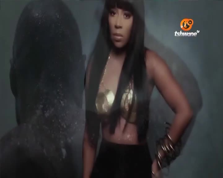 A woman in a gold top and black skirt stands with her arm bent, wearing multiple bangles. A dark, indistinct figure is to her left, with what looks like water droplets on its surface. A woman in a gold top and black skirt stands with her arm bent, wearing multiple bangles. A dark, indistinct figure is to her left, with what looks like water droplets on its surface.