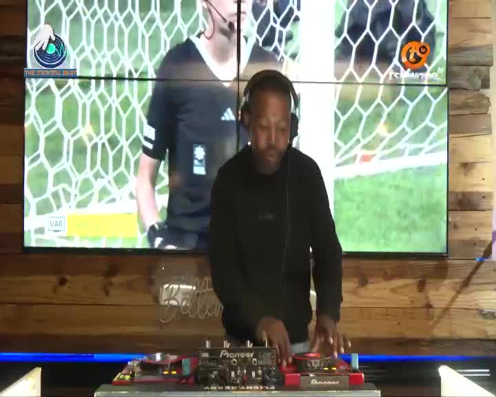 A DJ with headphones on works his equipment, his hands moving over the turntables. Behind him, a large screen shows a soccer referee, the Tshwane TV logo visible in the corner. A DJ with headphones on works his equipment, his hands moving over the turntables. Behind him, a large screen shows a soccer referee, the Tshwane TV logo visible in the corner.