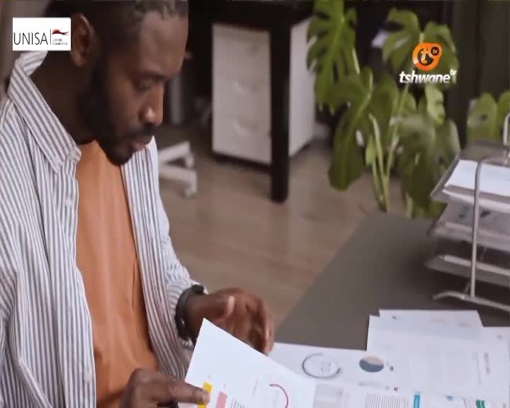 A man in a striped shirt leans over charts, his finger pointing at a section of a document. The Tshwane TV logo appears on a large plant in the background.