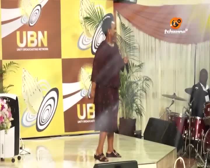 A woman in a maroon dress stands on a stage, speaking into a microphone. Behind her, a banner displays "UBN UNITY BROADCASTING NETWORK" and the Tshwane TV logo. A drummer plays in the background.