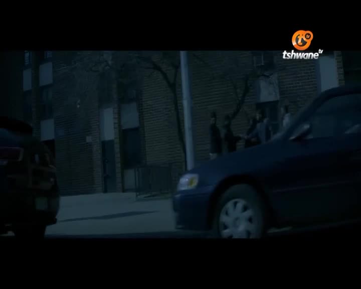 A dark blue sedan glides past a group of people gathered near a brick building. Another dark vehicle sits parked on the left, its shape barely visible in the dim light.