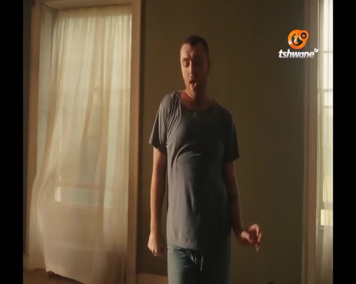 A man in a grey t-shirt and jeans moves his arms as he sings, his mouth open. The sheer curtains by the window flutter gently.