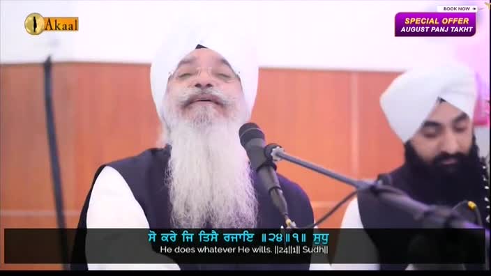 A man with a long white beard speaks into a microphone, his face lit by the studio lights. The Akaal Channel logo is visible in the corner, and another man sits beside him, also wearing a turban.
