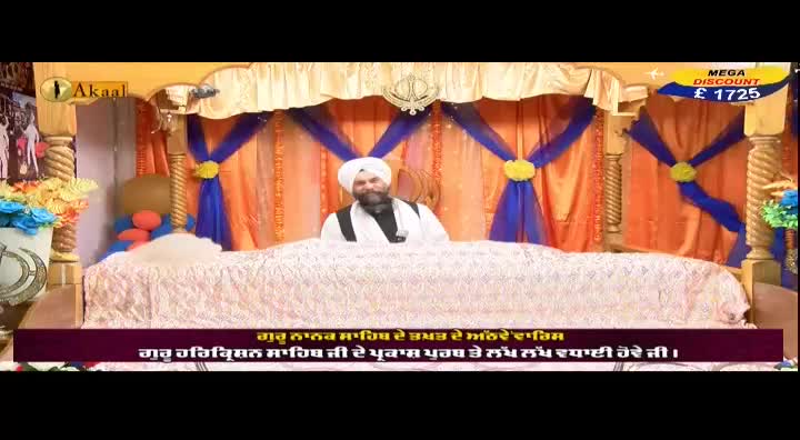 A man in a white turban and robes sits behind a large, ornate cloth-covered structure, likely in a religious setting. The Akaal Channel logo is visible in the corner, and the scene is decorated with colorful drapes.
