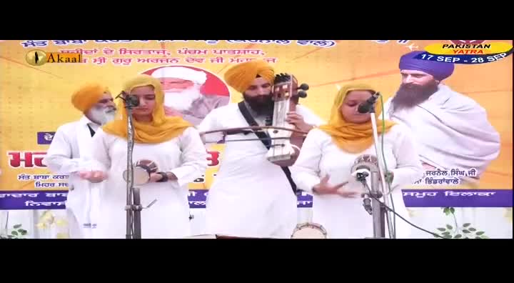 A group of people in white robes and turbans are performing, with two women in yellow headscarves holding small drums. A man with a stringed instrument stands in the center, and a banner for "Pakistan Yatra" is visible in the background.
