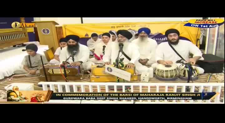 A group of men in white robes and turbans are seated, some playing instruments, others speaking into microphones. The scene is being broadcast on Akaal Channel from a Gurdwara in Handsworth, Birmingham, UK.
