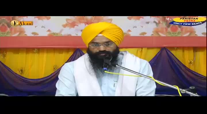 A man with a yellow turban and a long beard speaks into a microphone. Behind him, a backdrop of yellow and purple drapes hangs, and the Akaal Channel logo is visible.

