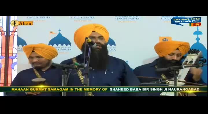A man with a long black beard speaks into a microphone, his voice broadcast on Akaal Channel. Two other men in turbans sit beside him, one playing a stringed instrument.
