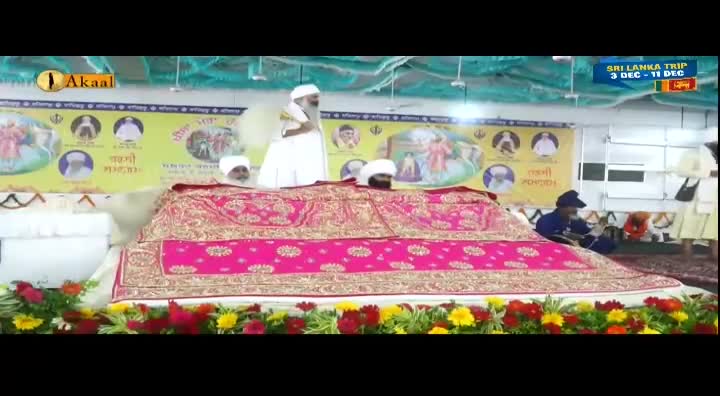 A man in white robes stands at a decorated platform, speaking. The scene is being broadcast on Akaal Channel, with a banner advertising a trip to Sri Lanka.
