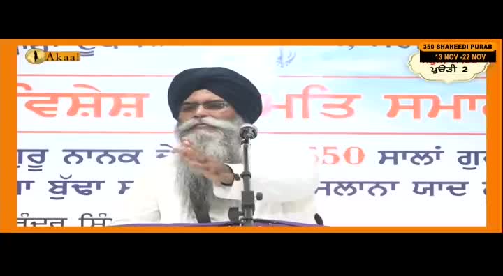 A man with a long white beard and a dark turban speaks into a microphone. Behind him, a banner advertises an event, likely broadcast on Akaal Channel from the United Kingdom.
