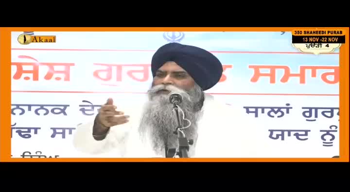 A man with a long white beard and a dark turban speaks into a microphone, gesturing with his hands. The Akaal Channel logo is visible, and a banner behind him displays text in Punjabi.
