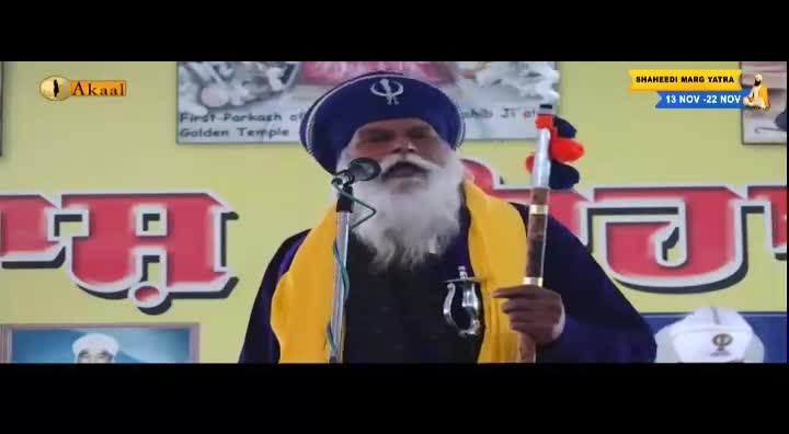 A man with a long white beard speaks into a microphone, holding a staff with red and orange tassels. The Akaal Channel logo is visible, and a banner announces the Shaheedi Marg Yatra.
