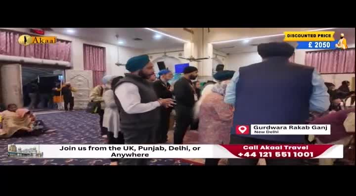 People in turbans and vests are gathered inside a building, some facing the camera, others looking towards the entrance. A banner for Akaal Travel, with a phone number, is visible at the bottom of the screen.
