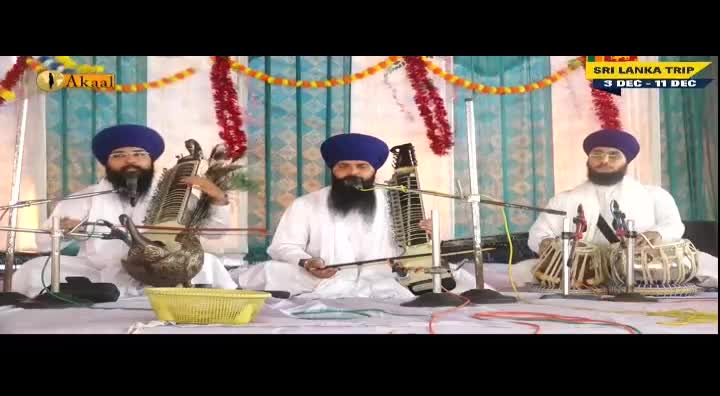 Three men in white robes and blue turbans sit cross-legged, playing instruments on a stage, broadcast by Akaal Channel. The central figure draws a bow across a stringed instrument, while the others pluck and tap their instruments in unison.
