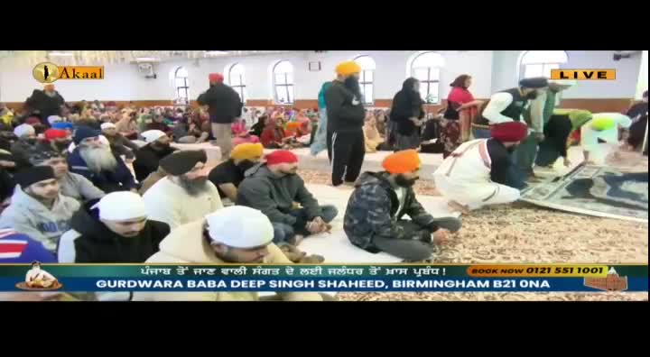 People sit and kneel on the floor in a large hall, many wearing turbans. The Akaal Channel is broadcasting live from the Gurdwara Baba Deep Singh Shaheed in Birmingham.
