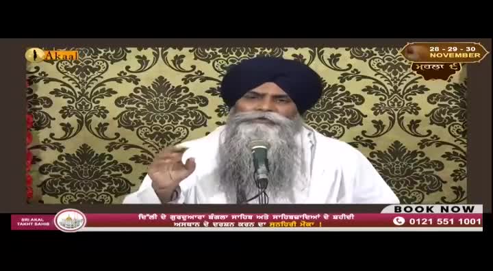 On the Akaal Channel, a man with a long, white beard speaks into a microphone. He gestures with his right hand, against a backdrop of a patterned wall.
