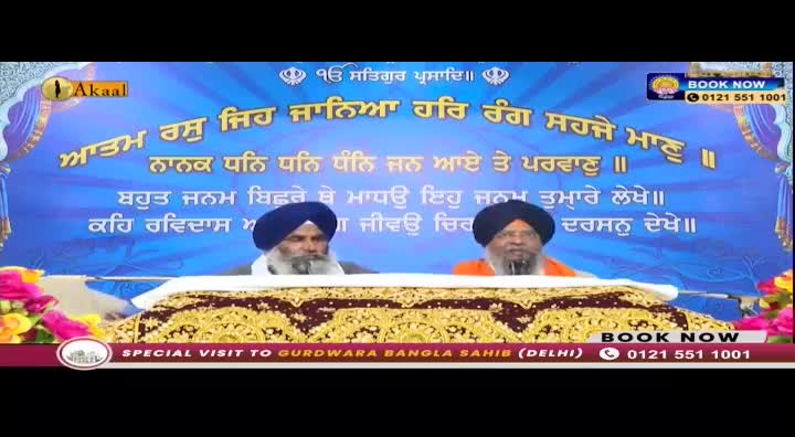 Two men in turbans sit behind a decorated cloth, speaking to the camera. A banner behind them displays text in Punjabi, with the Akaal logo visible.