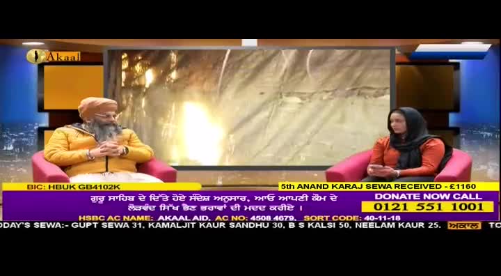 A man with a long beard, wearing a yellow jacket and turban, sits in a pink armchair. Across from him, a woman in an orange top and black headscarf holds a phone. A screen behind them displays a bright, abstract image.