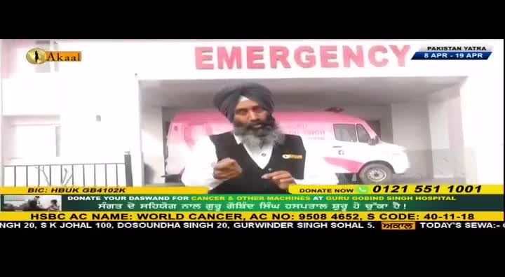 A man in a turban speaks in front of an emergency room entrance. A pink and white ambulance is parked to his right.